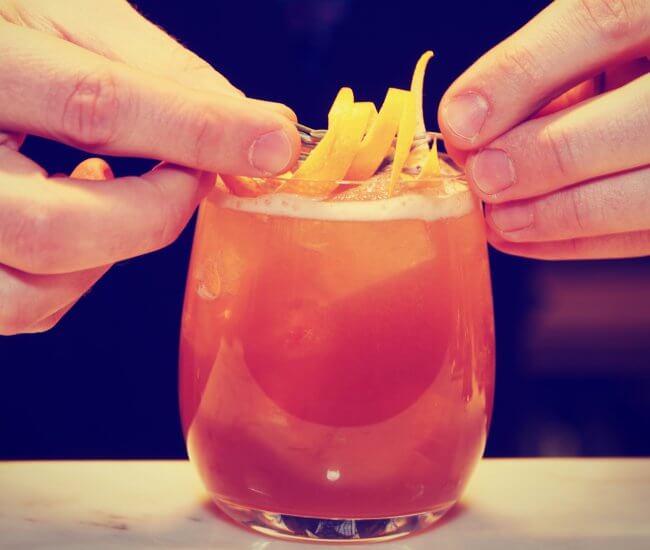 color photo of a close up of two hands placing lemon peel garnish on top of a red cocktail