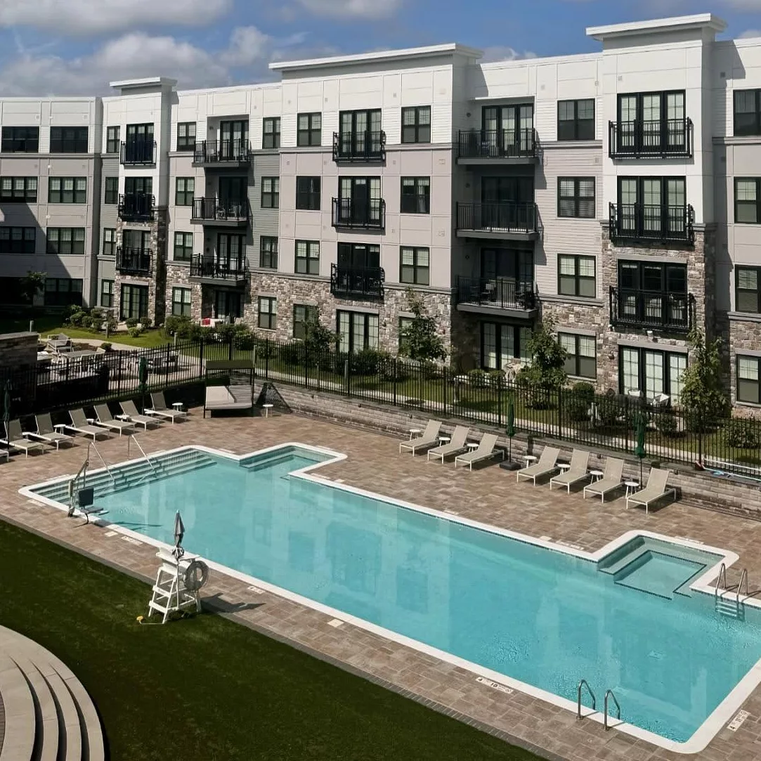 A pool surrounded by a brick patio and lounge chairs with an apartment building in the background and a blue sky with clouds.